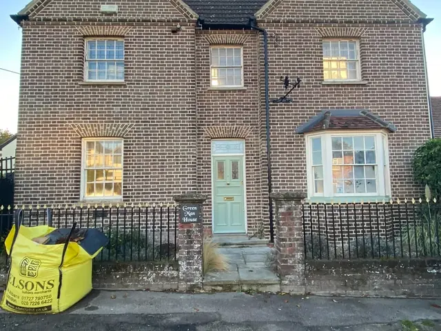 Large period brick house viewed from the street before masonry work, with a waste bag outside.