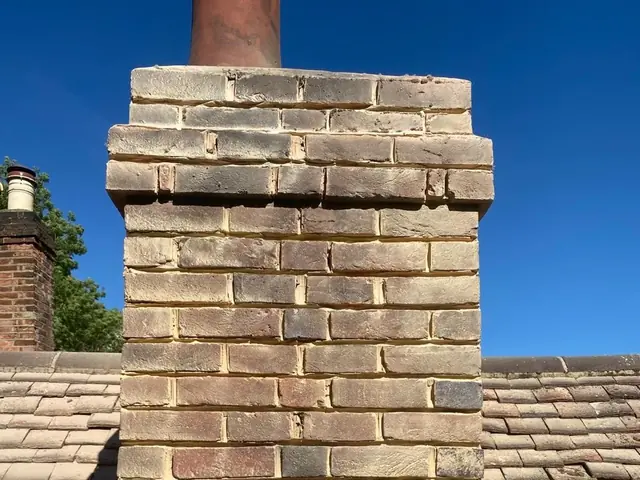 Repointed brick chimney stack on a tiled roof beneath a blue sky.