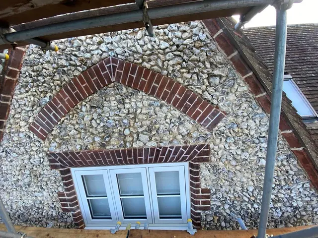 Repaired flint wall around a white framed arched window.